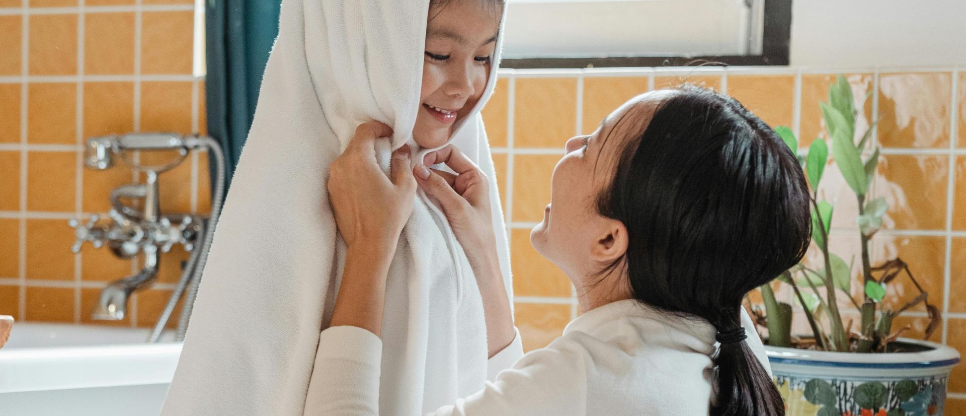 Cheerful ethnic mother wiping happy little daughter with large white towel in cozy light bathroom